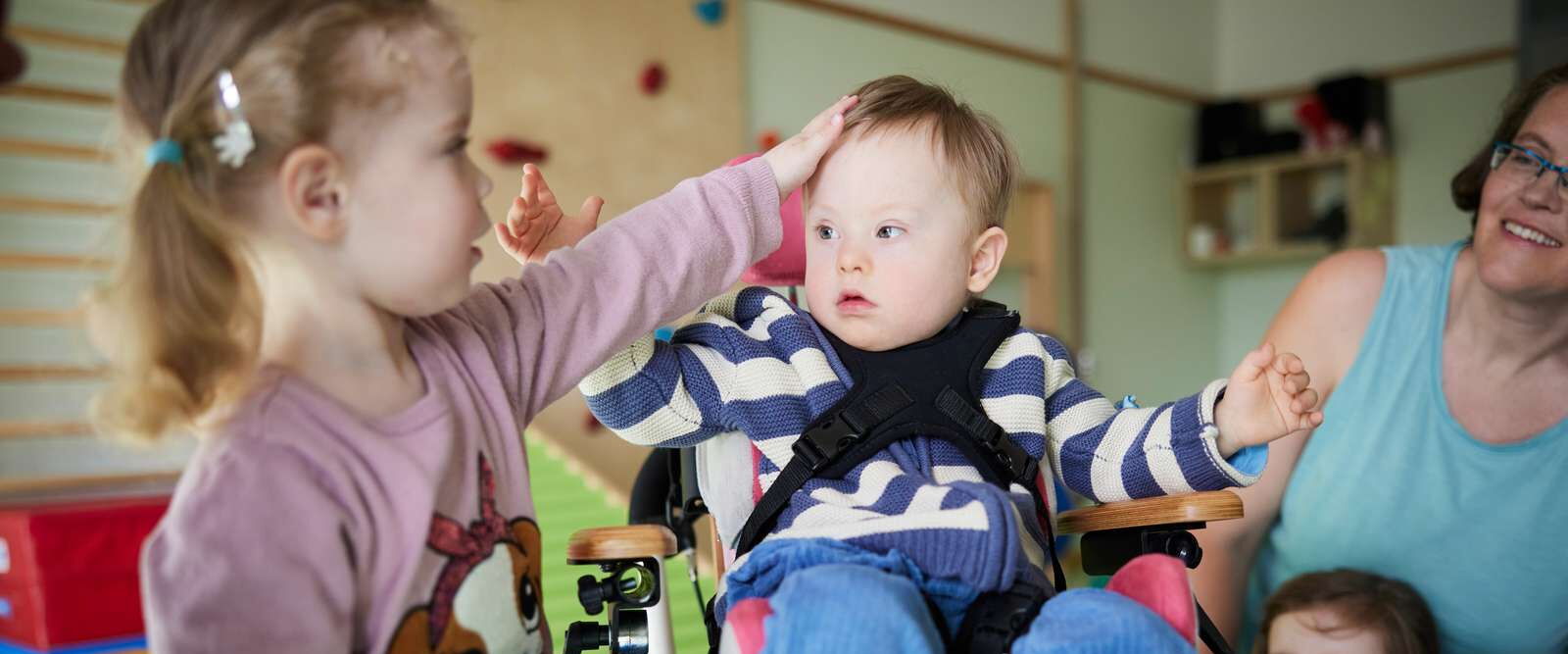 Ein kleines Mädchen streichelt einem kleinen Jungen in einem Stützstuhl über den Kopf, im Hintergrund spielen weitere Kinder
